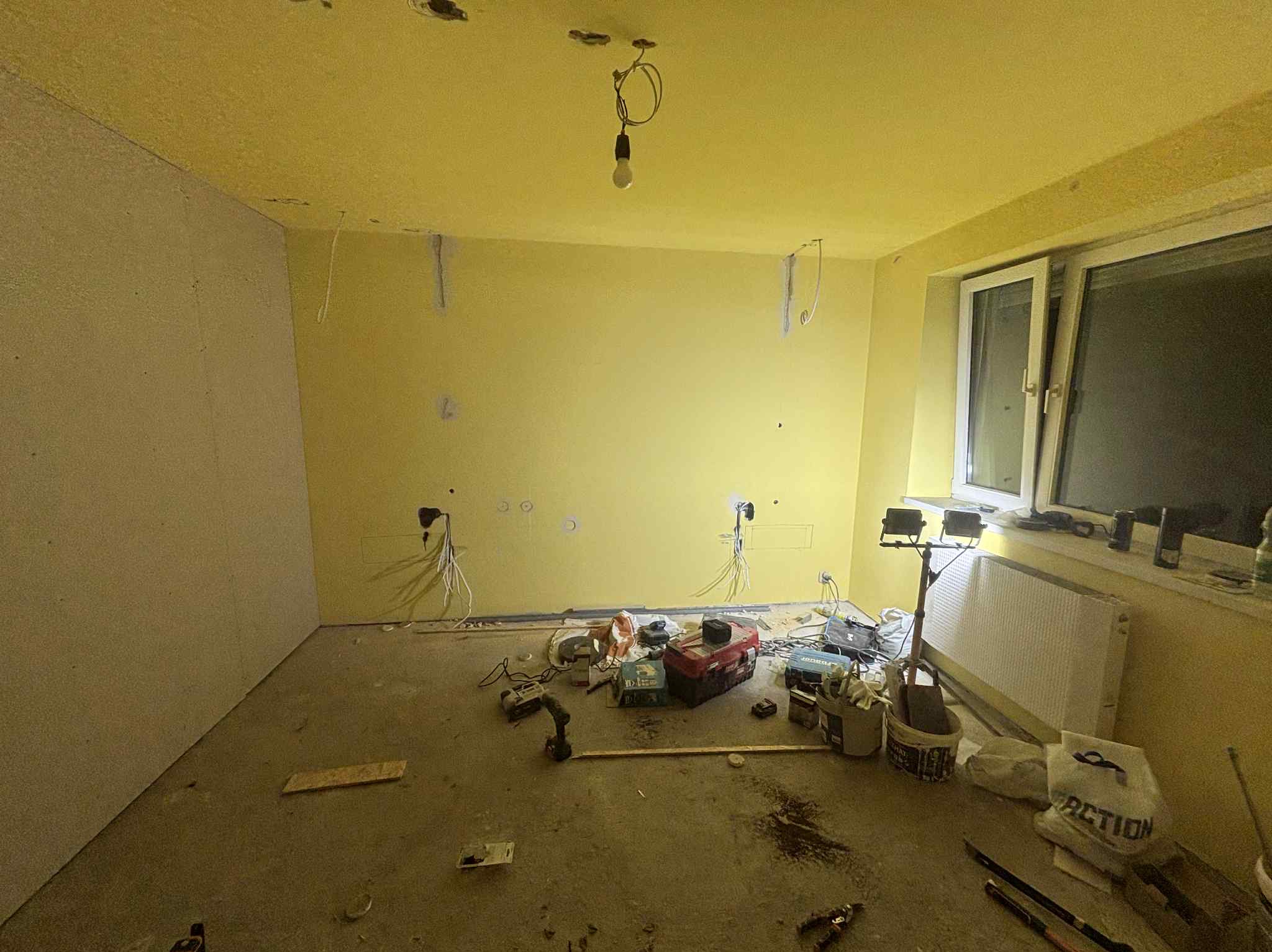 Pokój w trakcie remontu: ściany częściowo z płyt gipsowych, żółte ściany, widoczne kable, narzędzia i akcesoria budowlane. Żarówka zwisa z sufitu.