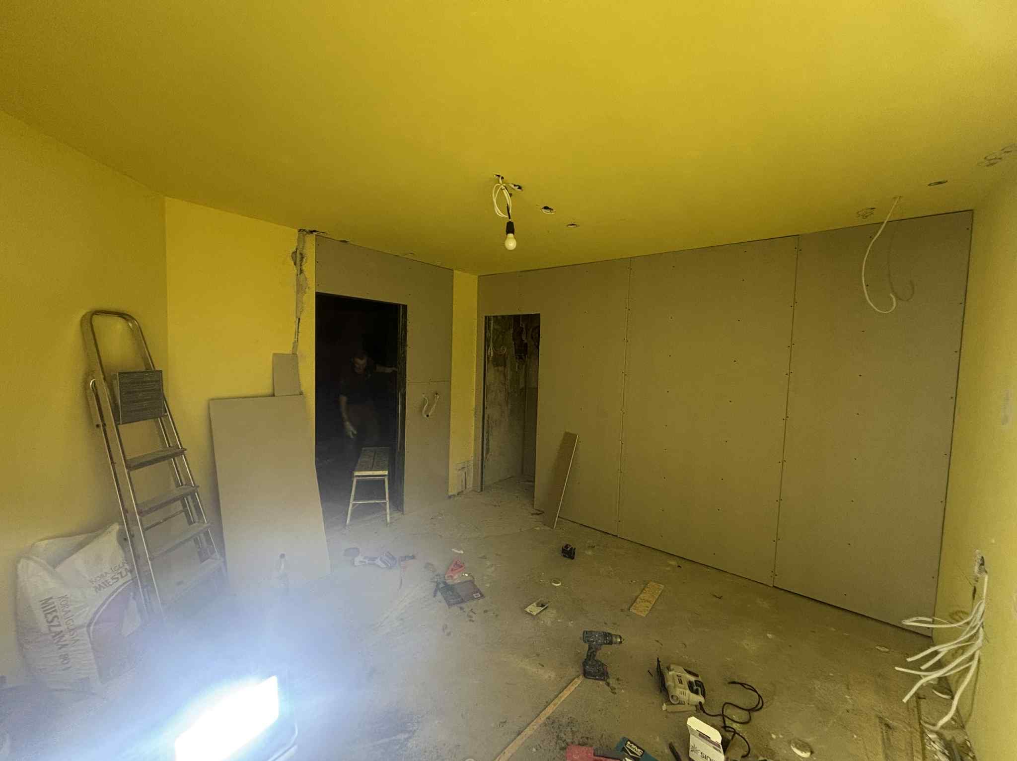 Pokój w trakcie remontu: ściany z płyt karton-gips, widoczne narzędzia, drabina oparta o ścianę. Żółte ściany i sufit. W tle widoczna postać w przejściu.