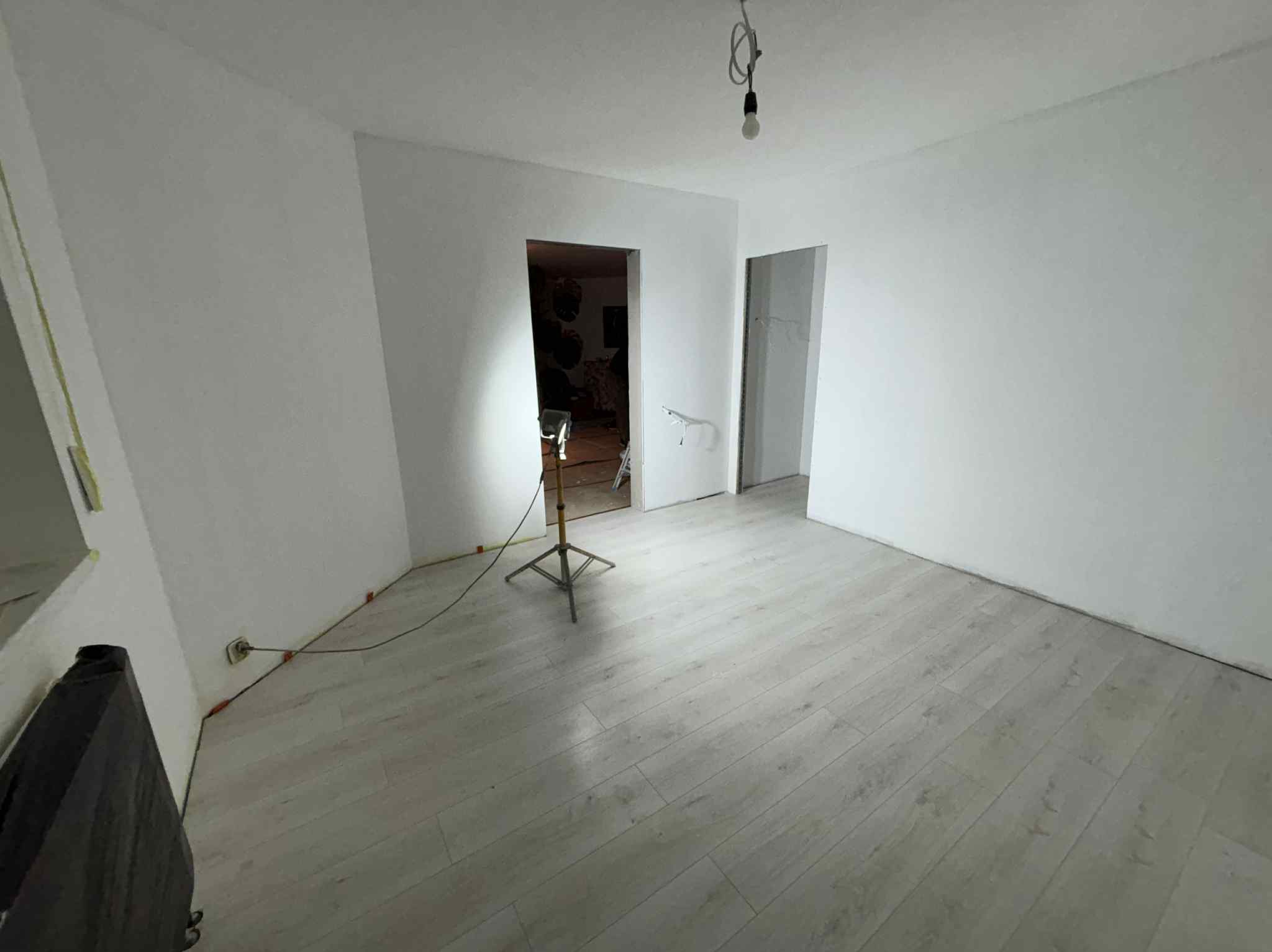 Jasne panele podłogowe w wyremontowanym pokoju z białymi ścianami, oświetlonym lampą budowlaną. Widoczne gniazdka elektryczne i kable.