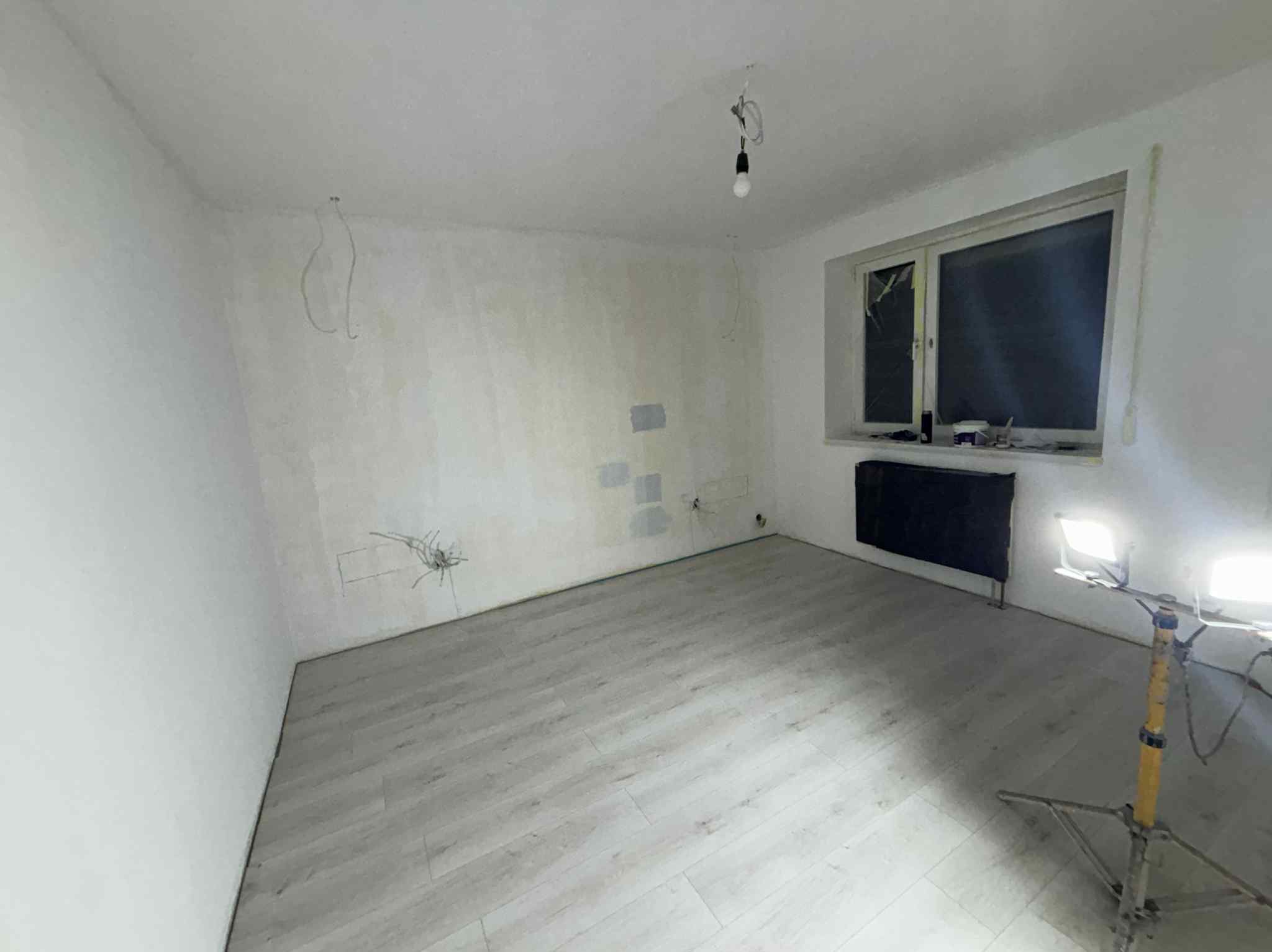 Pokój po szpachlowaniu i ułożeniu paneli, widoczne niedociągnięcia na ścianach, lampa budowlana oświetla wnętrze, okno z parapetem.