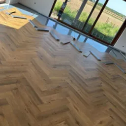 Układanie parkietu w jodełkę na podkładzie w nowym domu z dużym oknem. Widoczne panele i narzędzia. Część podłogi już wykończona. Słoneczny dzień.