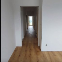 Bartłomiej Miśkiewicz - Korytarz po remoncie z ułożonymi panelami podłogowymi w odcieniach brązu i świeżo pomalowanymi ścianami na biało. Widok w głąb korytarza, wpadające światło dzienne.