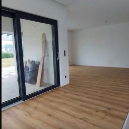 Bartłomiej Miśkiewicz - Jasna podłoga z paneli ułożona w przestronnym wnętrzu z dużym przesuwnym oknem. Widoczne materiały budowlane sugerują etap wykończeniowy.