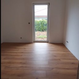 Bartłomiej Miśkiewicz - Jasne panele podłogowe w pokoju z białymi ścianami. Widok na zewnątrz przez przeszklone drzwi balkonowe. Minimalistyczne wykończenie wnętrza.