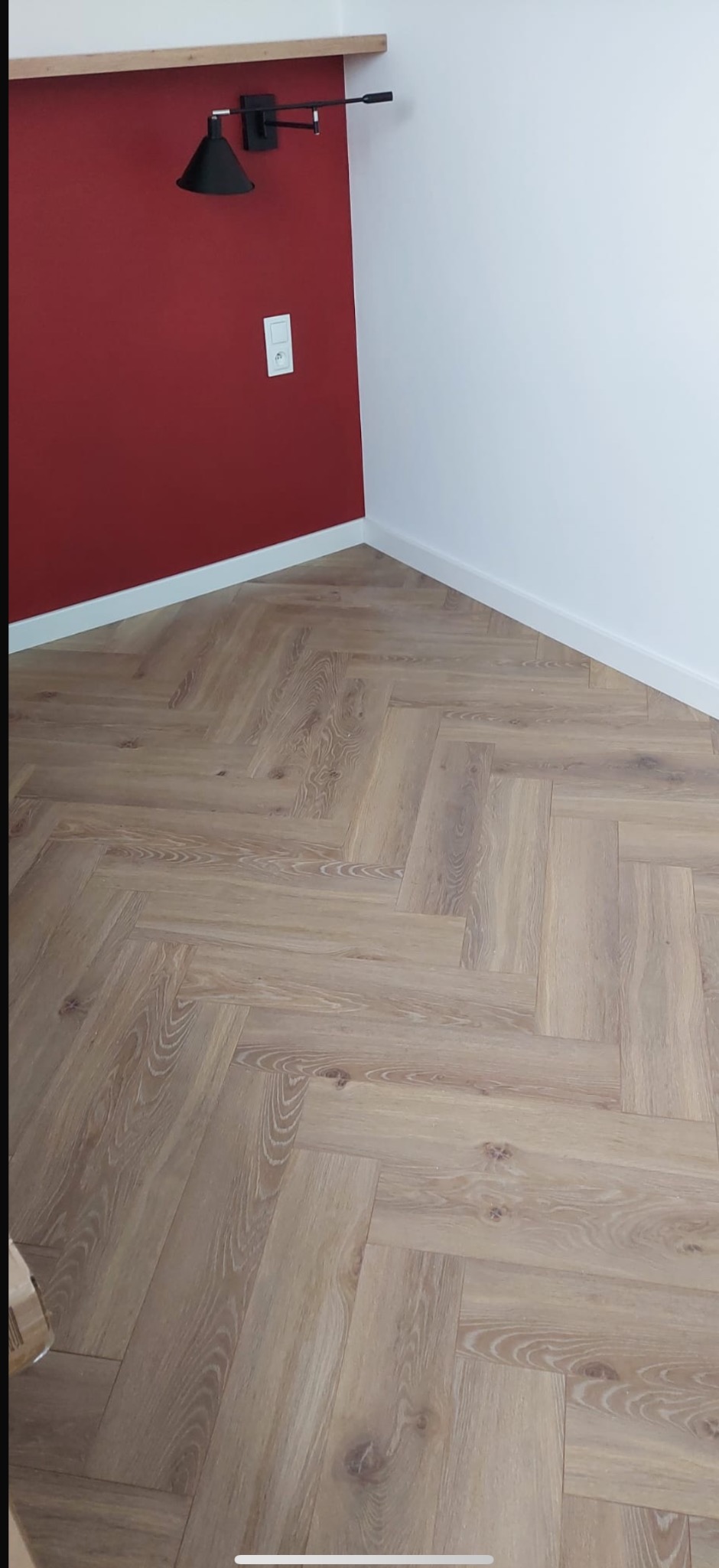 Jasny parkiet ułożony we wzór jodełki francuskiej w rogu pokoju z czerwoną i białą ścianą. Przy czerwonej ścianie czarna lampa.