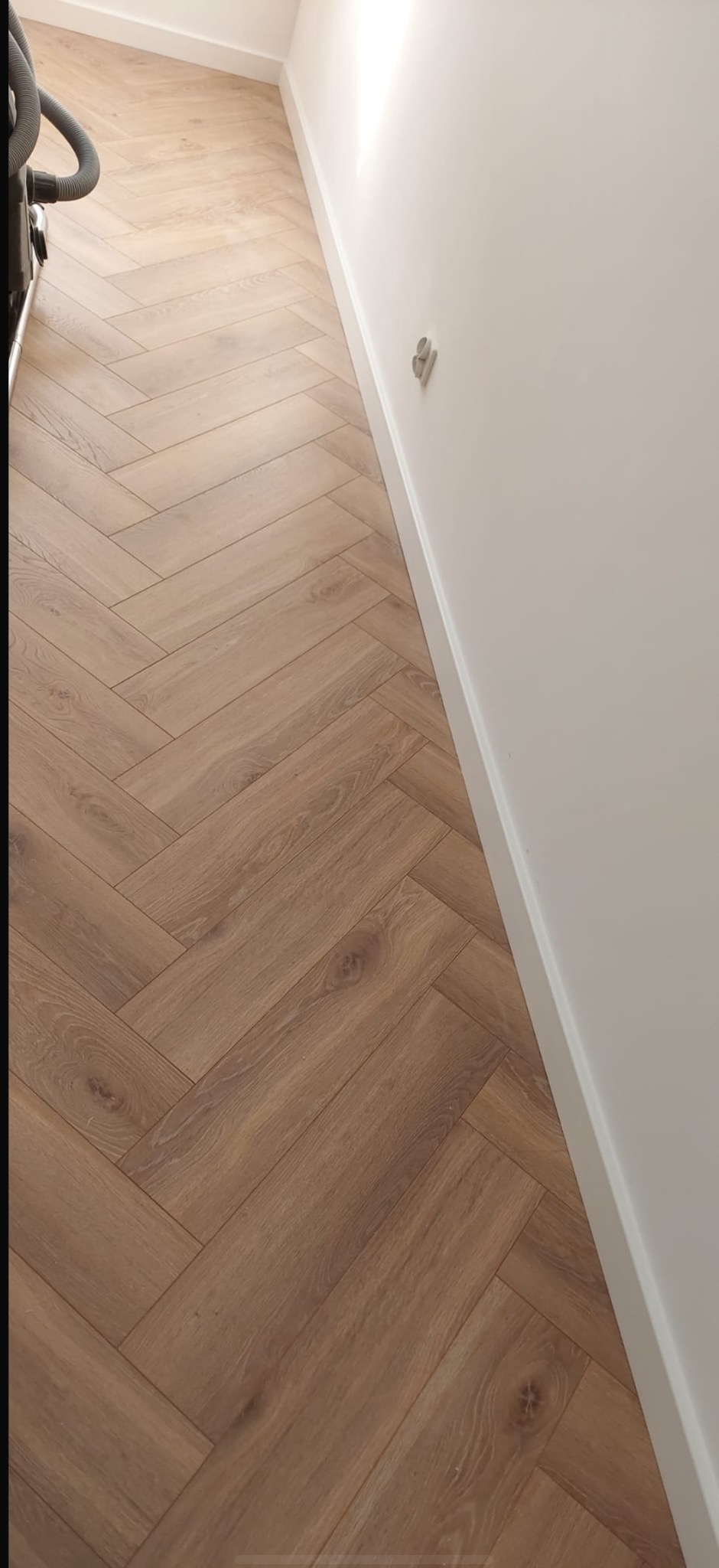 Ułożony parkiet w jodełkę francuską, widoczny fragment odkurzacza. Biała ściana z listwą przypodłogową i gniazdkiem. Naturalne światło, ciepłe odcienie drewna.