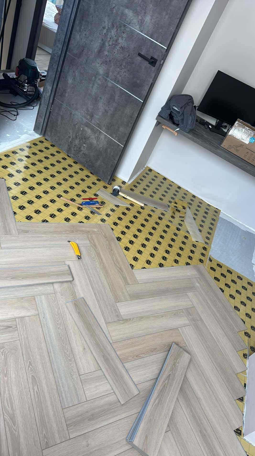 Układanie paneli podłogowych w jodełkę na podkładzie, widoczne narzędzia i fragment szarej ściany z drzwiami. Częściowo ułożona podłoga.