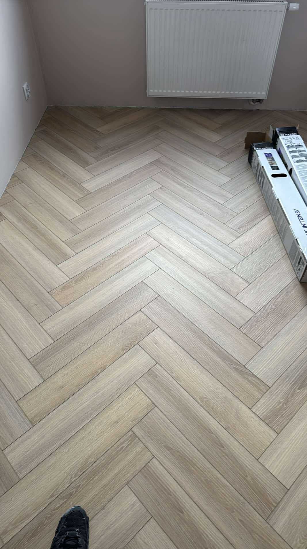 Ułożony parkiet w jodełkę, jasne drewno, widoczny grzejnik i opakowania paneli. Fragment buta w lewym dolnym rogu kadru. Naturalne światło dzienne.
