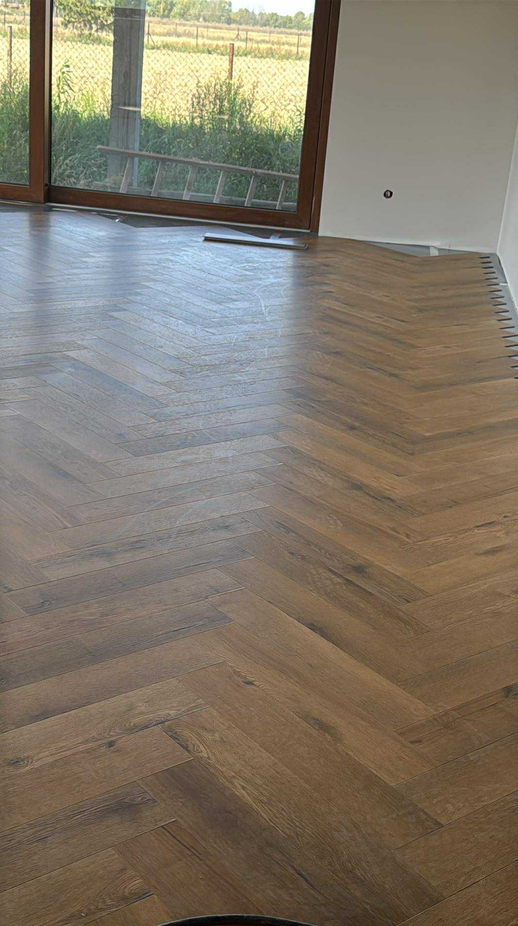 Ułożony parkiet w jodełkę, brązowe deski, widoczne listwy przypodłogowe, duże okno z widokiem na zewnątrz. Jasne wnętrze w trakcie wykończenia.