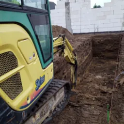 Żółta koparka Yanmar SV15 w trakcie prac ziemnych pod fundamenty w Pniewie. W tle ściany z białych bloczków.