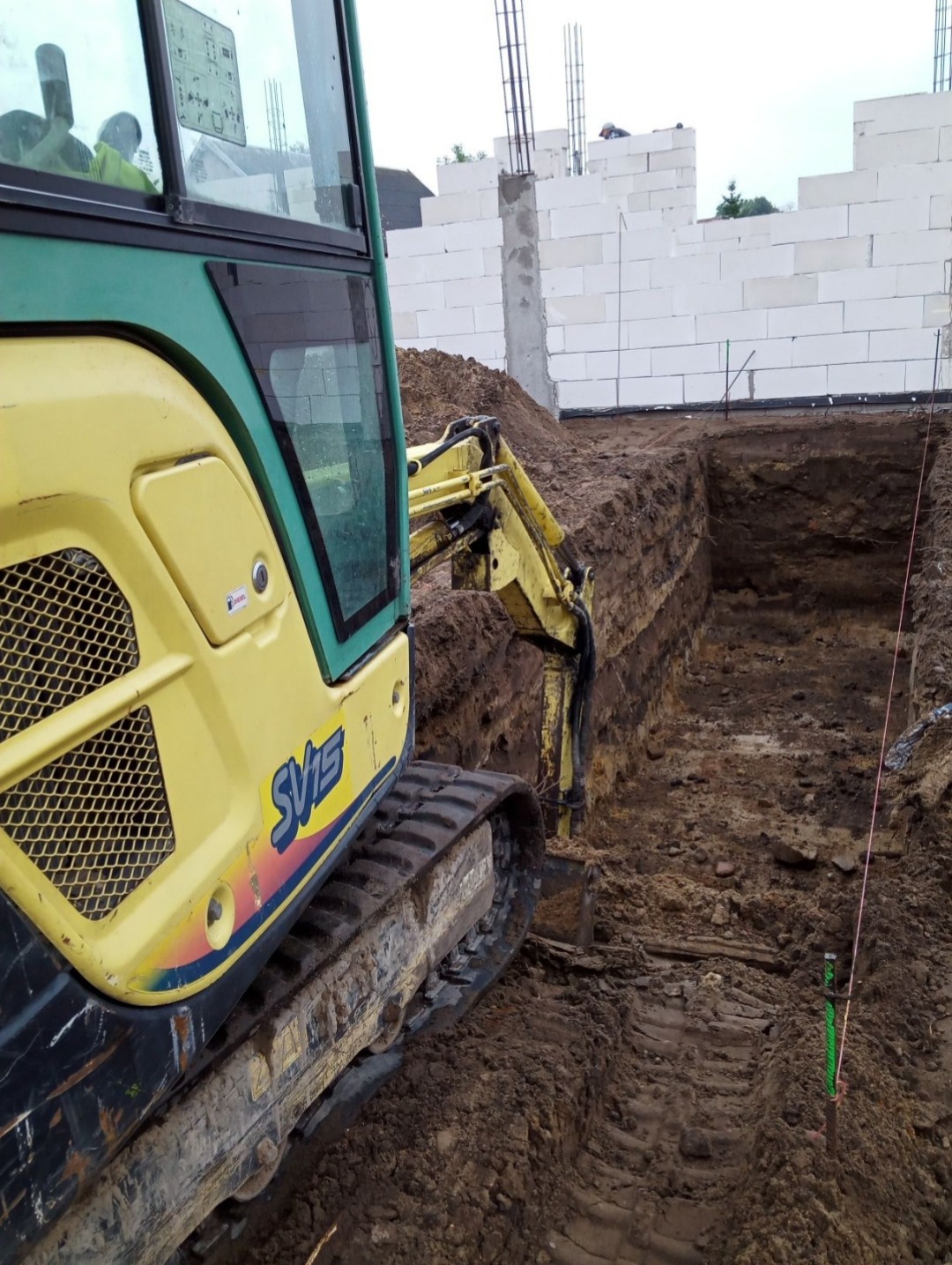 Żółta koparka Yanmar SV15 w trakcie prac ziemnych pod fundamenty w Pniewie. W tle ściany z białych bloczków.