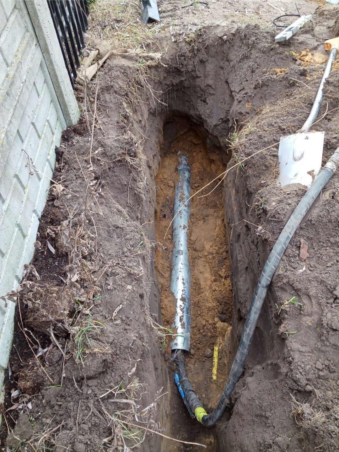Wykop w ziemi z rurą w osłonie i kablem. Widoczna łopata i ogrodzenie z prefabrykatów betonowych. Wykonanie instalacji podziemnej.