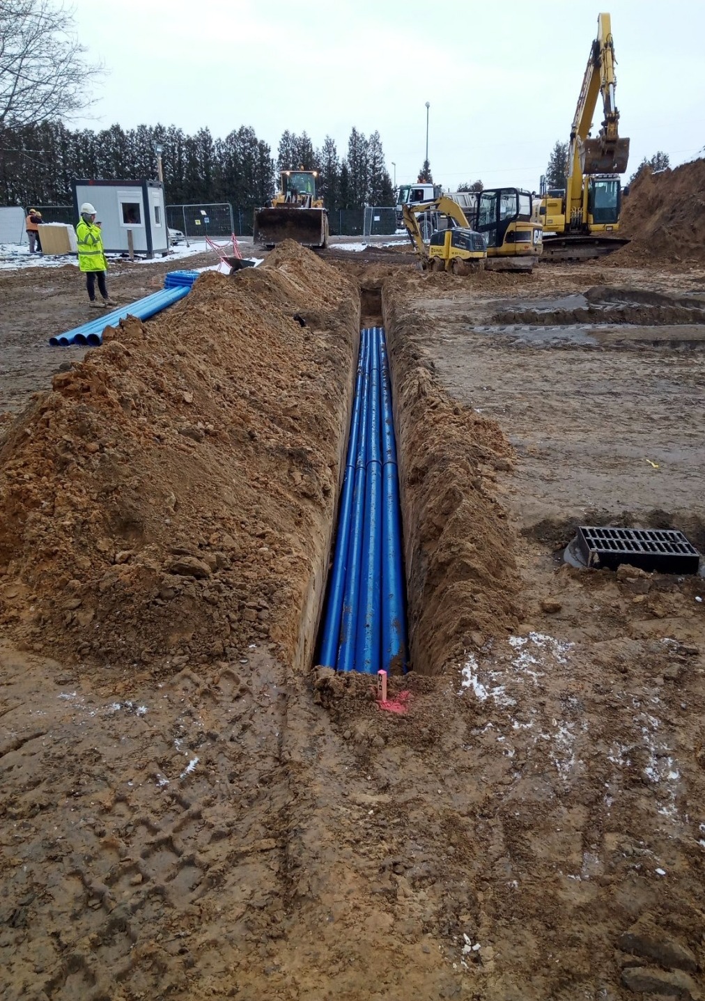 Wkop z ułożonymi niebieskimi rurami, widoczne maszyny budowlane i pracownik w odblaskowej kamizelce. Teren budowy, ziemia, częściowo zaśnieżony teren.