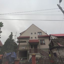 ANGEL HOUSE - Dom jednorodzinny w trakcie prac elewacyjnych, widoczne rusztowania i pracownicy. Szara, pochmurna pogoda. Ujęcie z dołu, zza ogrodzenia.