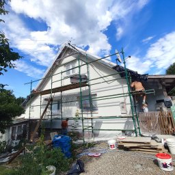 ANGEL HOUSE - Dom jednorodzinny w trakcie prac elewacyjnych. Widoczne rusztowania, pracownicy, wiaderka z farbą i zabezpieczone okna folią malarską. Błękitne niebo z chmurami w tle.