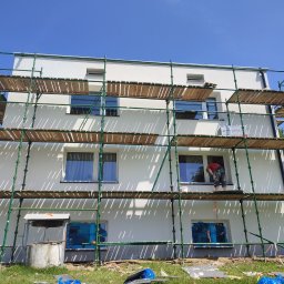 ANGEL HOUSE - Biała elewacja budynku w trakcie prac wykończeniowych. Widoczne rusztowania, zabezpieczone okna folią i pracownik w oknie. Słoneczny dzień.