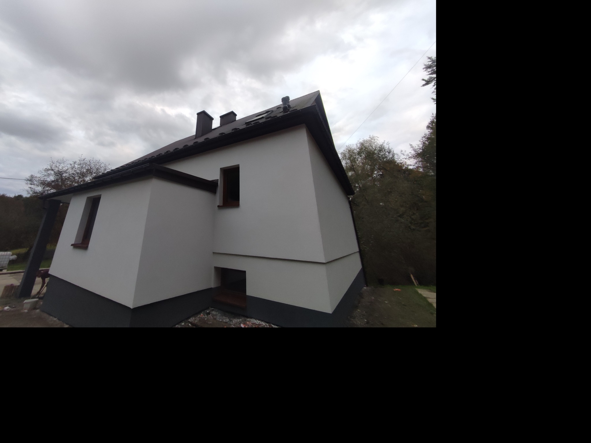 Boczny widok domu z nową, jasną elewacją i ciemną podmurówką. Widoczne okna z brązowymi ramami, ciemny dach i fragment zalesionego otoczenia w pochmurny dzień.