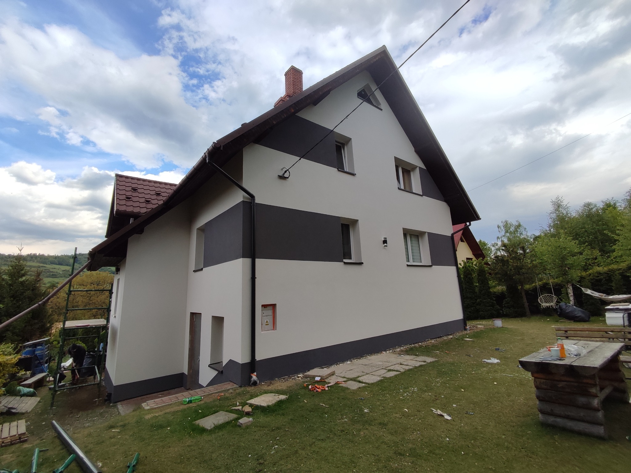 Dwukondygnacyjny dom z nową, dwukolorową elewacją (szary pas na białym tle) widziany z boku, z fragmentem rusztowania po lewej stronie i zielenią w tle.