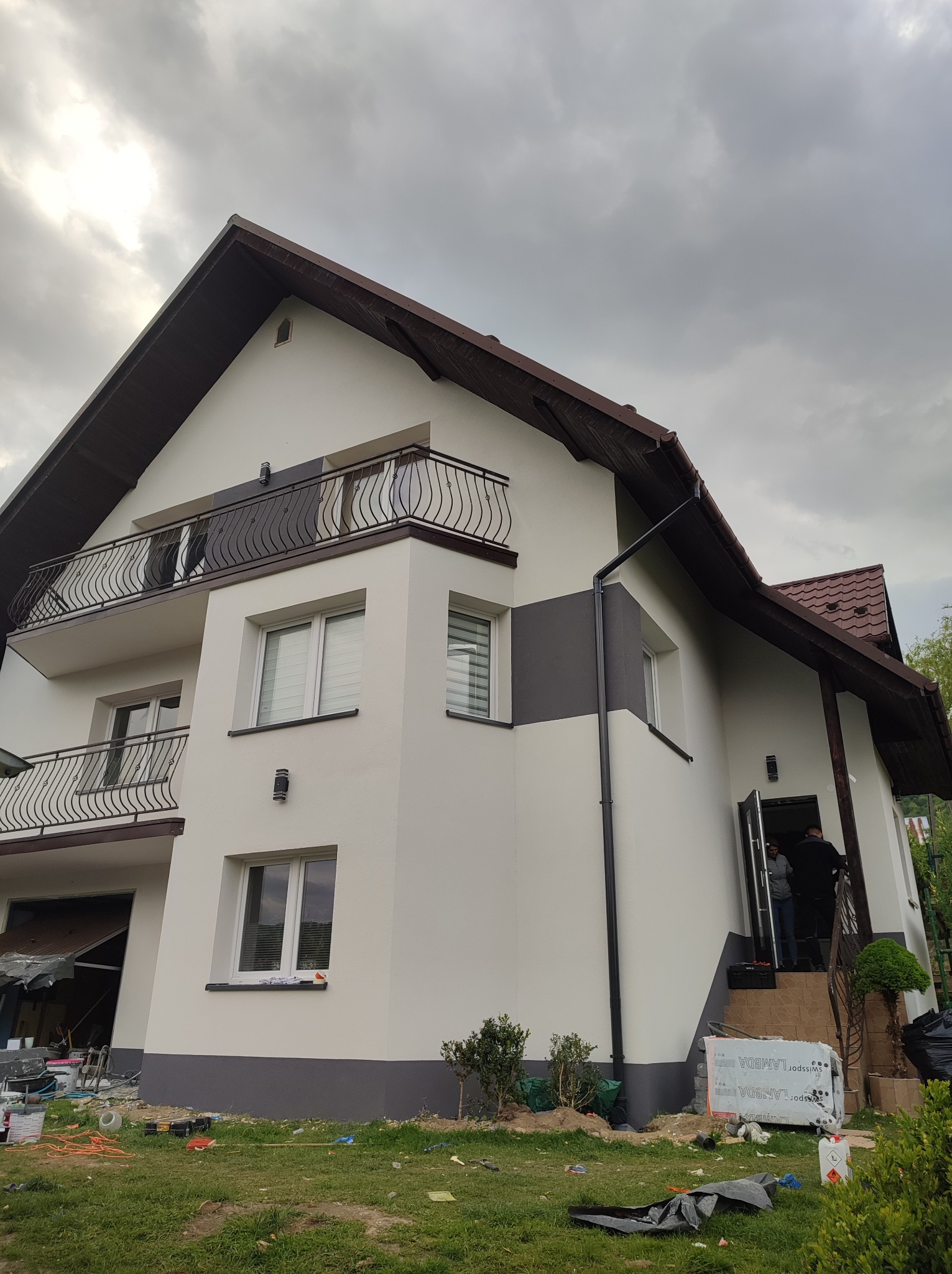 Dwupiętrowy dom z balkonami po renowacji elewacji, z widocznymi detalami wykończenia i elementami dekoracyjnymi. Ujęcie z dołu podkreśla bryłę budynku na tle pochmurnego nieba.