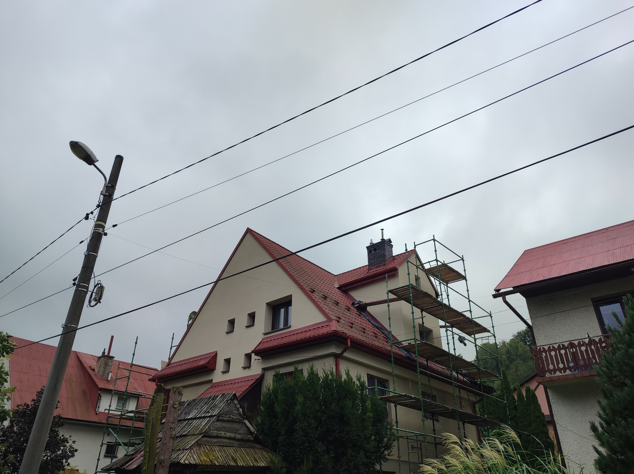 Dom z beżową elewacją i czerwoną dachówką w trakcie prac. Widoczne rusztowania, słup elektryczny i pochmurne niebo. Ujęcie z dołu.