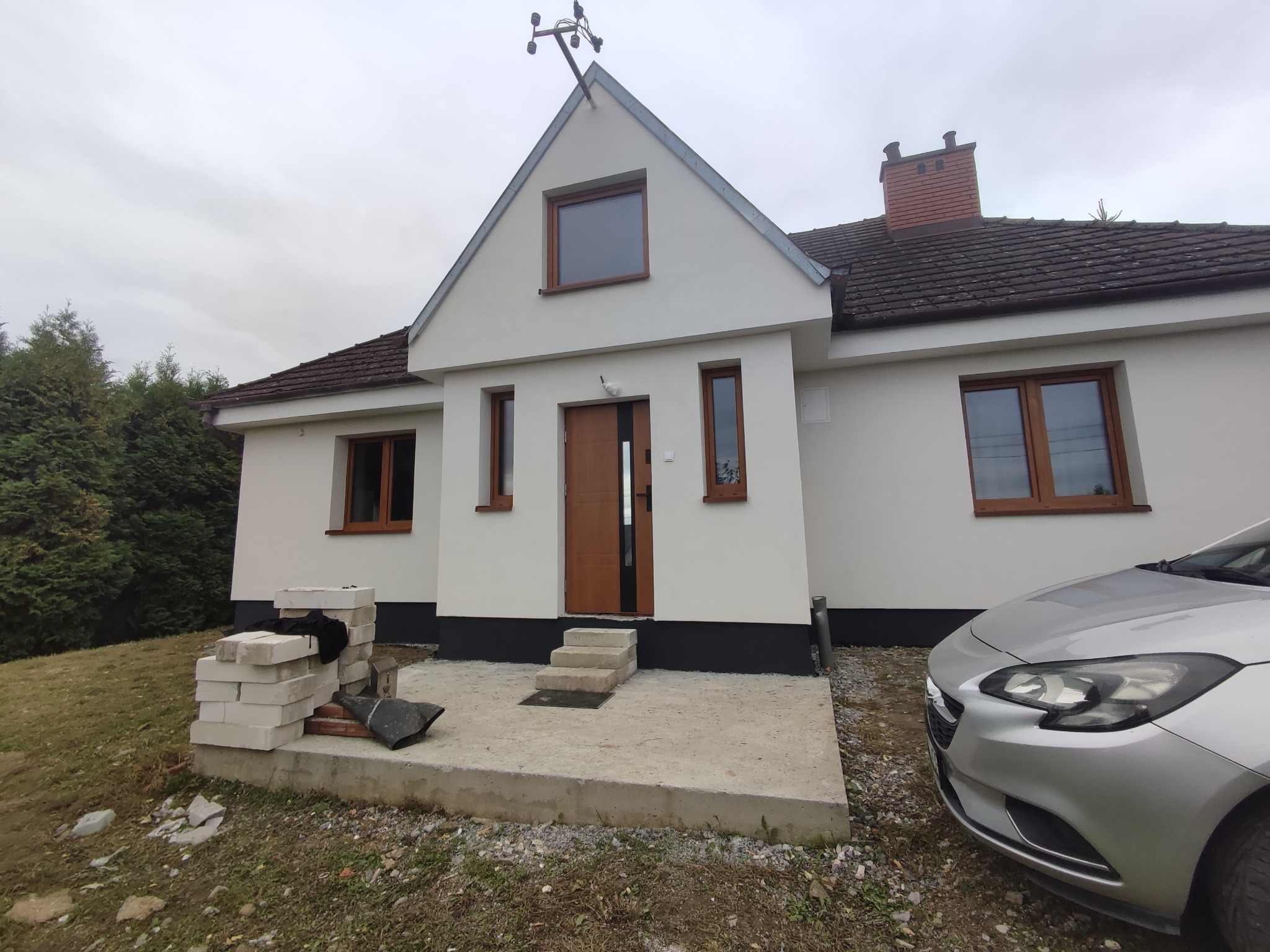 Dom jednorodzinny po odświeżeniu elewacji, z widocznymi blokami budowlanymi i samochodem osobowym. Ujęcie z poziomu gruntu, pochmurne niebo w tle.