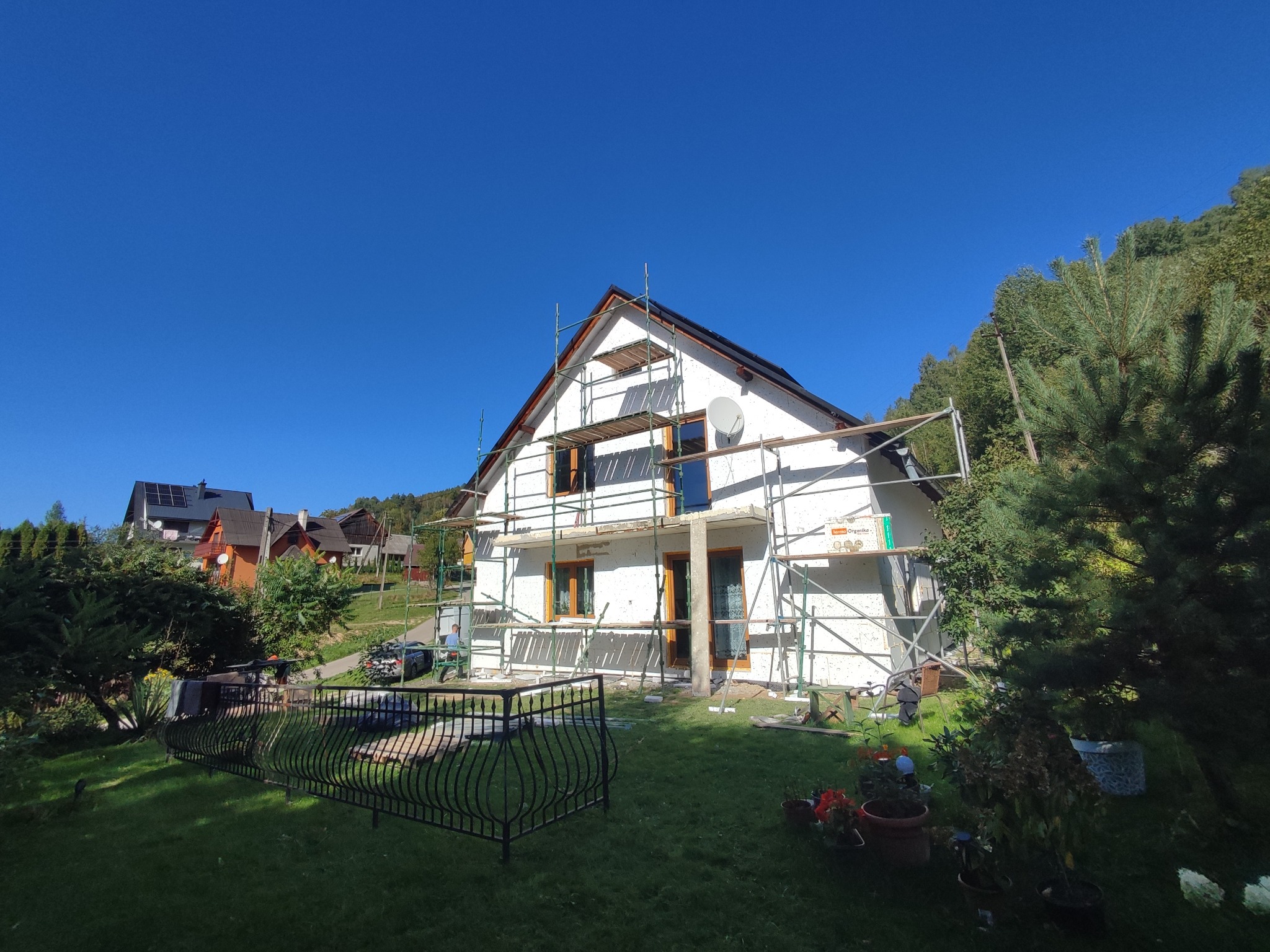 Biały dom jednorodzinny w trakcie ocieplania, otoczony rusztowaniami, na tle błękitnego nieba i zieleni. Widoczne detale: okna z drewnianą ramą, antena satelitarna.