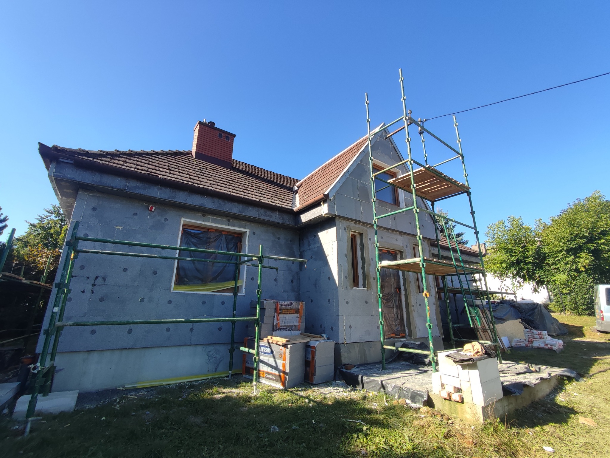 Dom jednorodzinny w trakcie ocieplania ścian zewnętrznych, widoczne rusztowania, styropian i materiały budowlane. Słoneczny dzień, błękitne niebo.