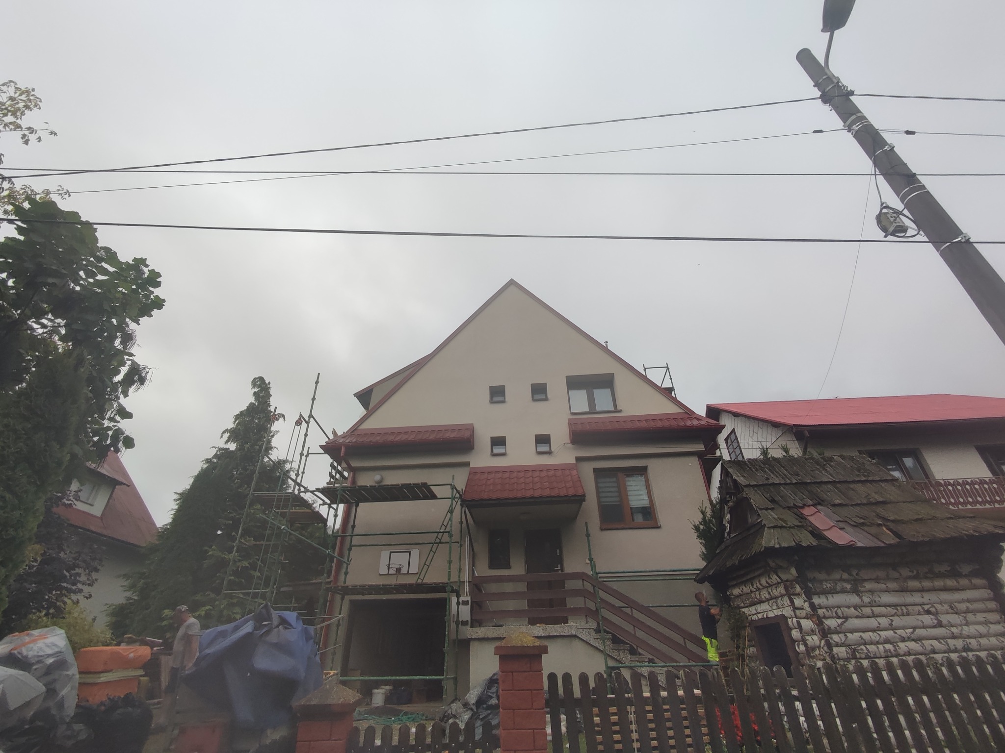 Dom jednorodzinny w trakcie prac elewacyjnych, widoczne rusztowania i pracownicy. Szara, pochmurna pogoda. Ujęcie z dołu, zza ogrodzenia.