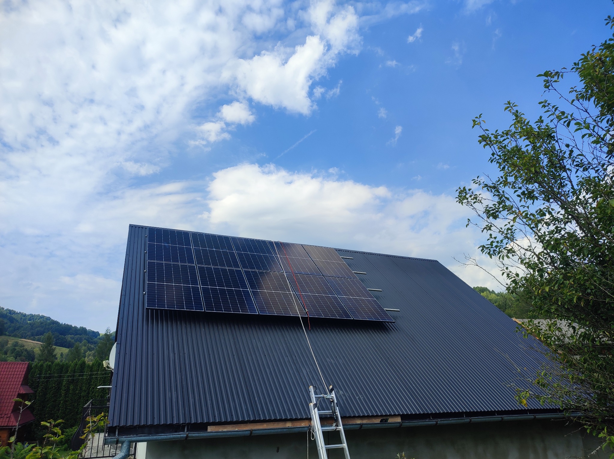 Ciemny dach z panelami słonecznymi w trakcie montażu, drabina oparta o dach, błękitne niebo z chmurami w tle. Widok z dołu, częściowo zasłonięty przez drzewo.