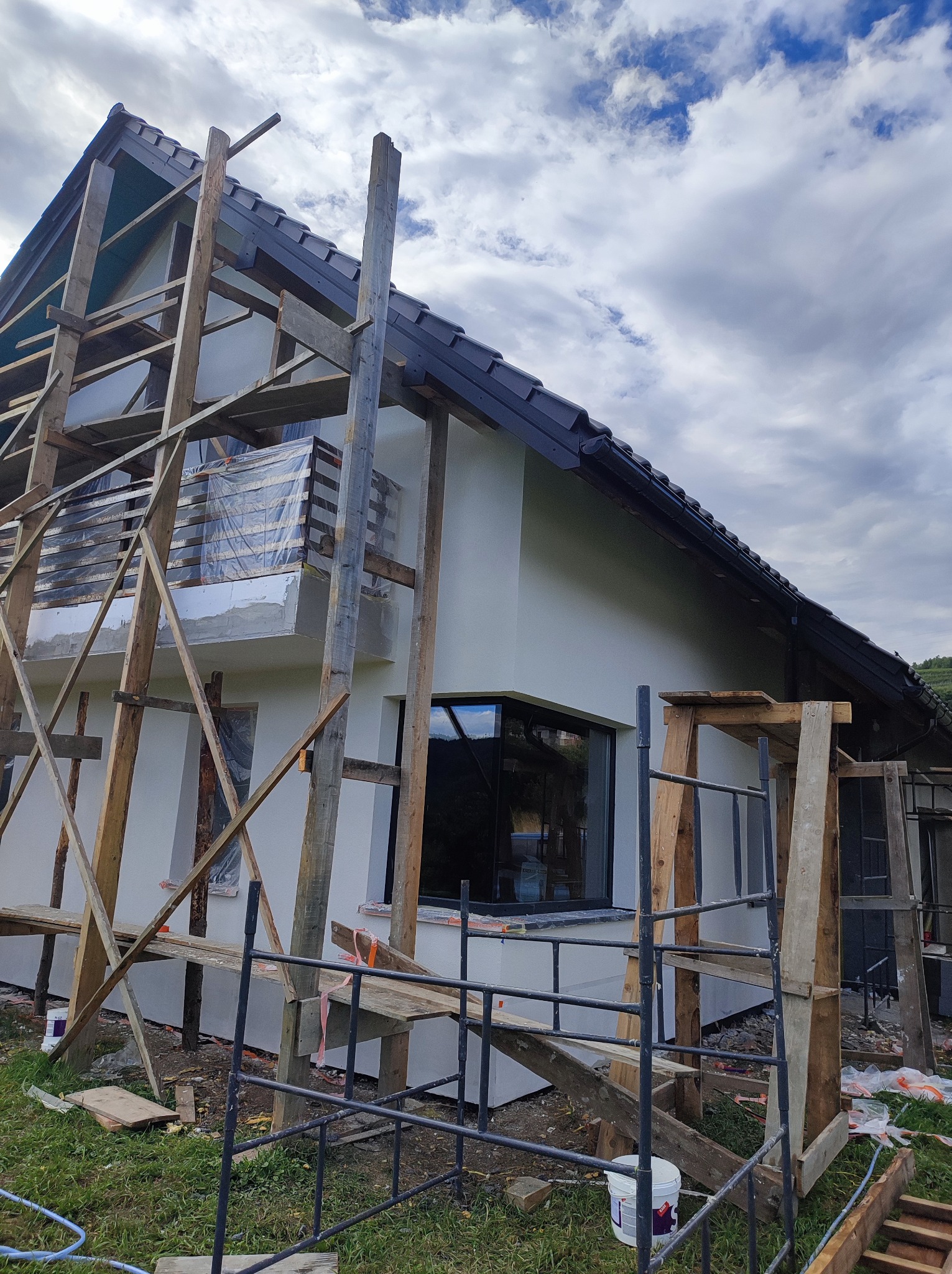 Biała elewacja domu jednorodzinnego w trakcie malowania, widoczne rusztowania drewniane i metalowe, okna z czarnymi ramami, pochmurne niebo w tle.