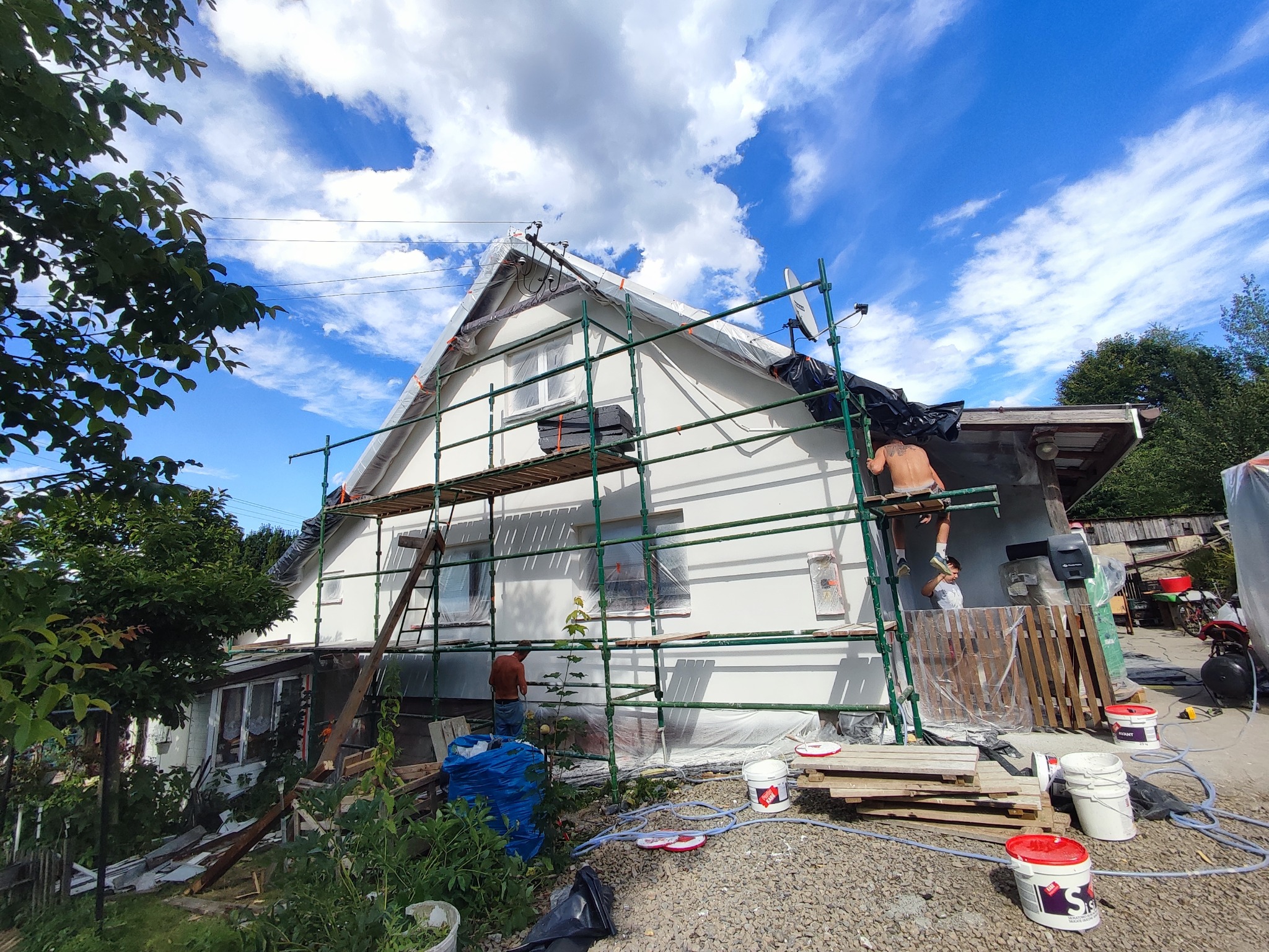 Dom jednorodzinny w trakcie prac elewacyjnych. Widoczne rusztowania, pracownicy, wiaderka z farbą i zabezpieczone okna folią malarską. Błękitne niebo z chmurami w tle.