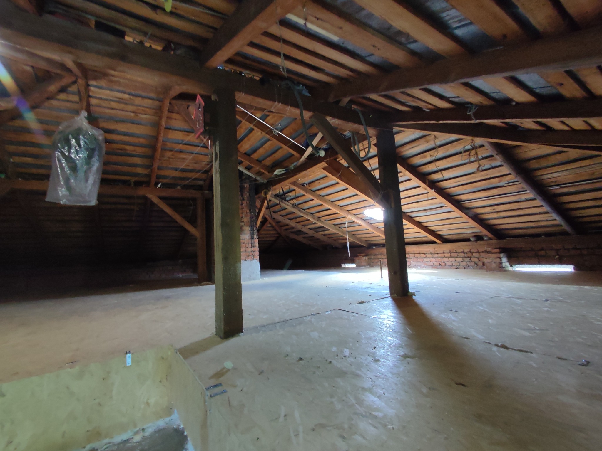 Poddasze z drewnianą konstrukcją dachu i oknem dachowym. Widoczne ceglane elementy i izolacja. Ujęcie z perspektywy podłogi, podkreślające przestrzeń i surowy charakter.