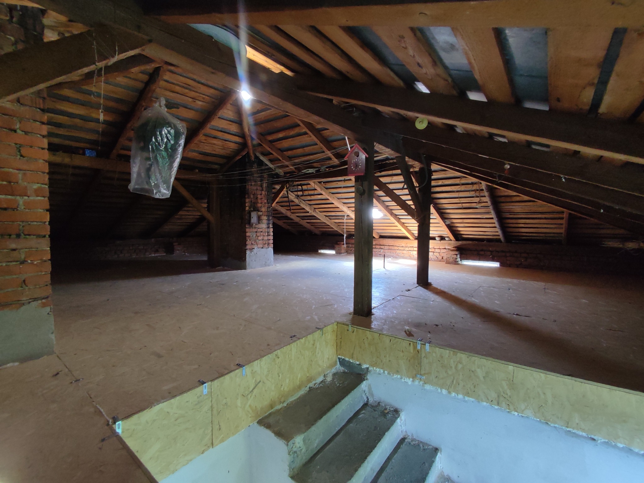 Poddasze z drewnianą konstrukcją dachu, oświetlone naturalnym światłem. Widoczne elementy muru z cegły, drewniane słupy i podłoga z płyt OSB. Częściowo wykończone schody.