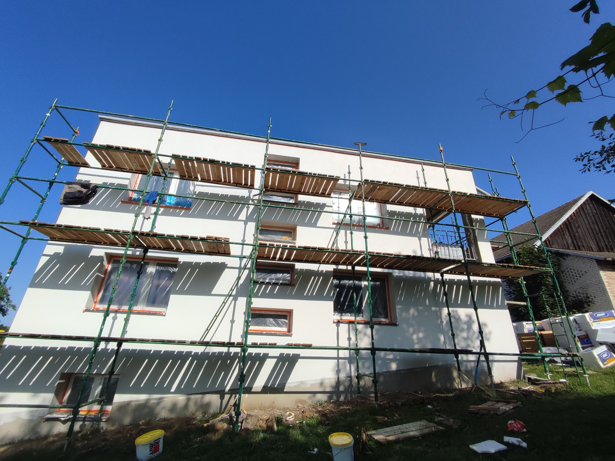 Biały dom w rusztowaniach, okna zabezpieczone folią. Widoczne puszki z farbą i styropian. Słoneczny dzień, błękitne niebo. Ujęcie z dołu, podkreślające wysokość budynku.
