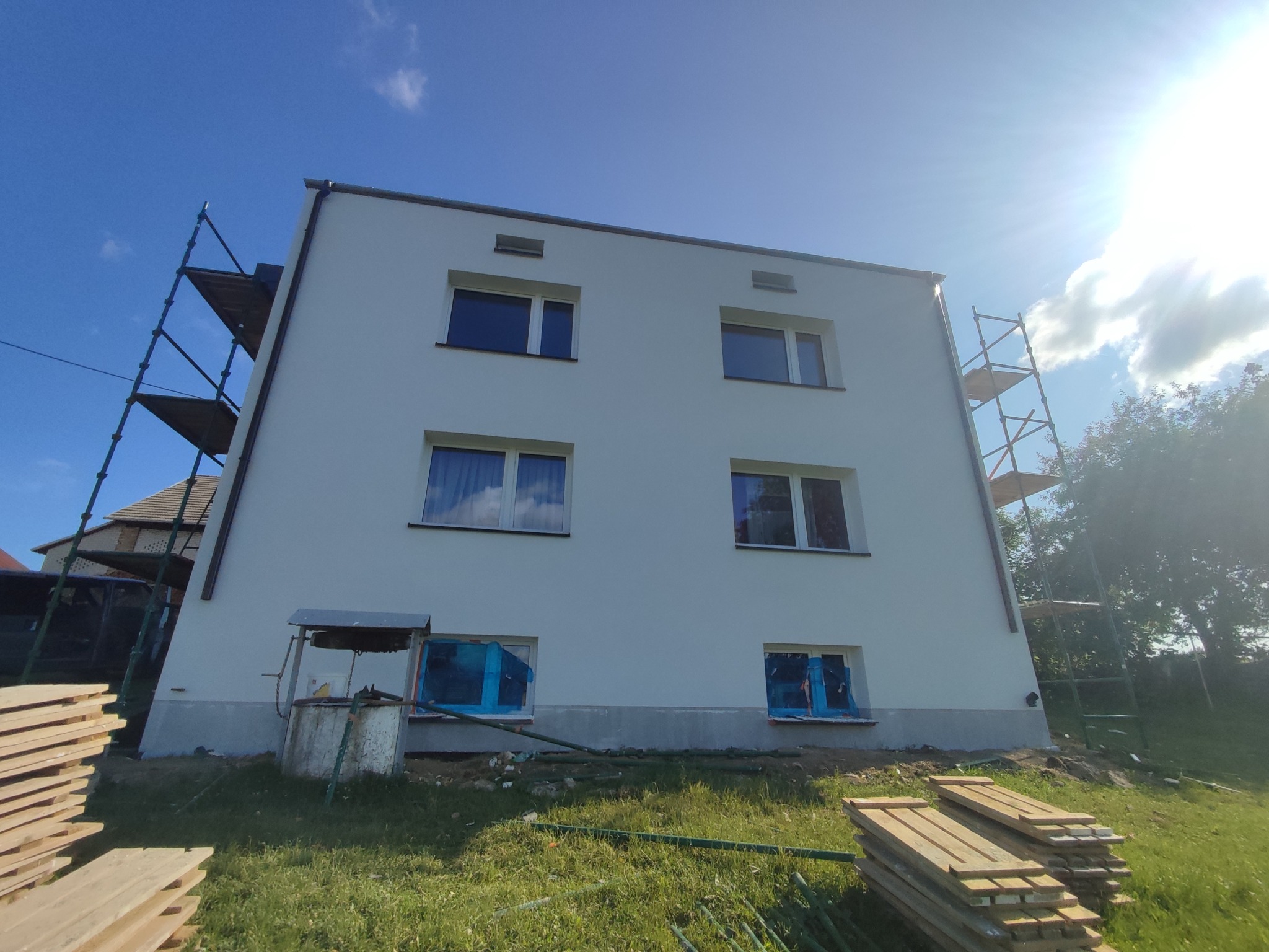 Dwupiętrowy dom po odnowieniu elewacji, z oknami zabezpieczonymi folią. Widoczne rusztowania i studnia. Słoneczny dzień, trawa i ułożone deski na pierwszym planie.