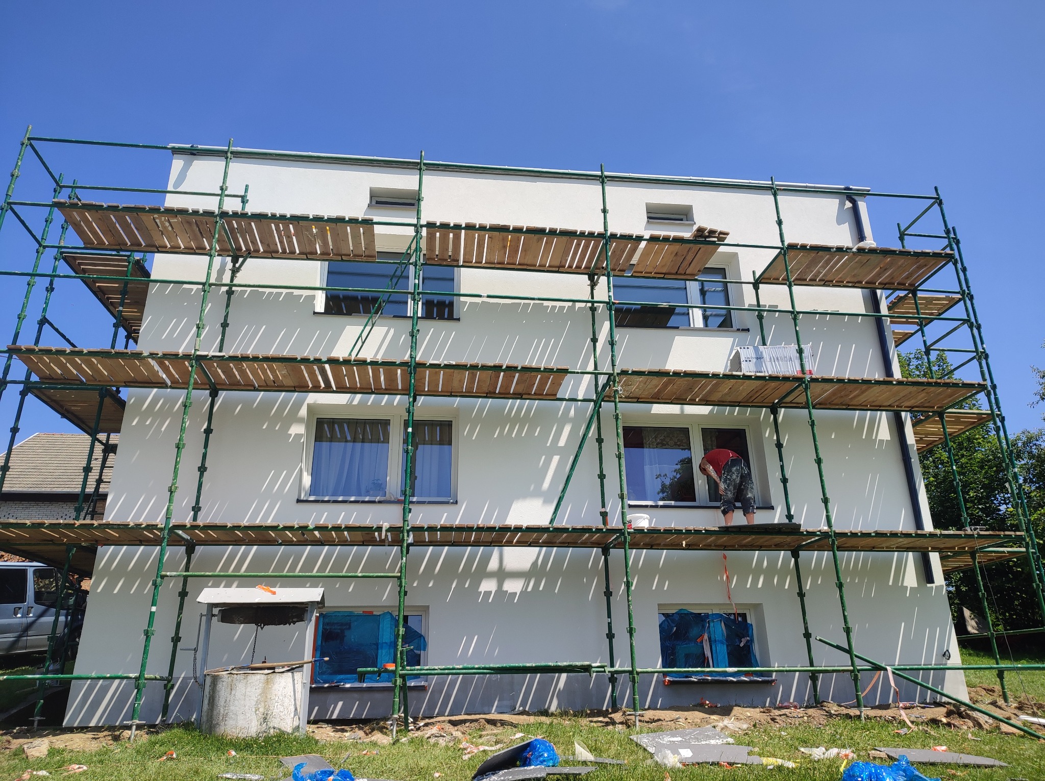 Biała elewacja budynku w trakcie prac wykończeniowych. Widoczne rusztowania, zabezpieczone okna folią i pracownik w oknie. Słoneczny dzień.