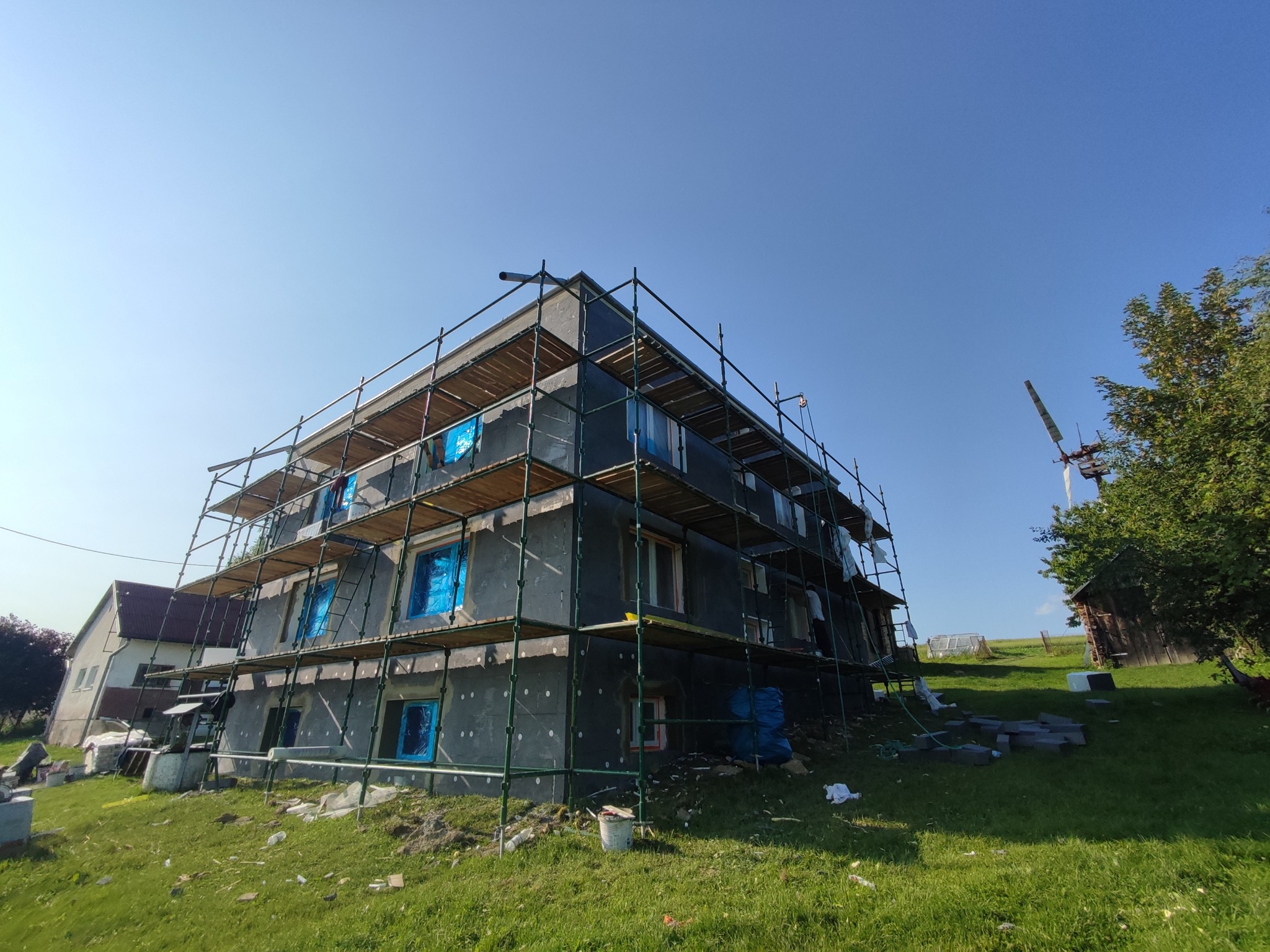 Budynek w trakcie ocieplania, otoczony rusztowaniem. Okna zabezpieczone folią. Widoczne płyty izolacyjne na ścianach. Prace w toku, na tle zieleni i błękitnego nieba.