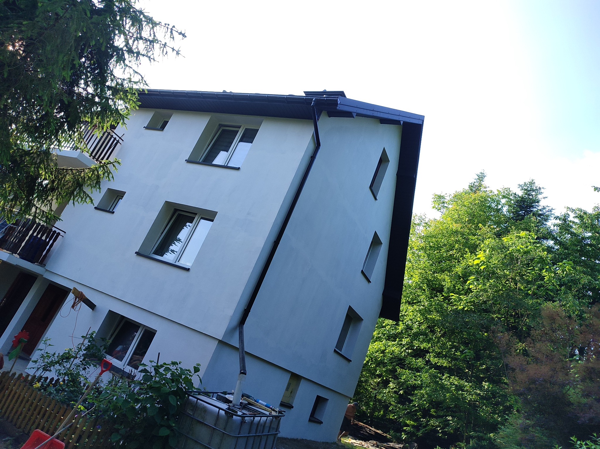 Jasny, dwupiętrowy dom z balkonem, po renowacji elewacji, widziany z dołu pod kątem. Widoczne okna, rynna i fragment ogrodu z zielenią.