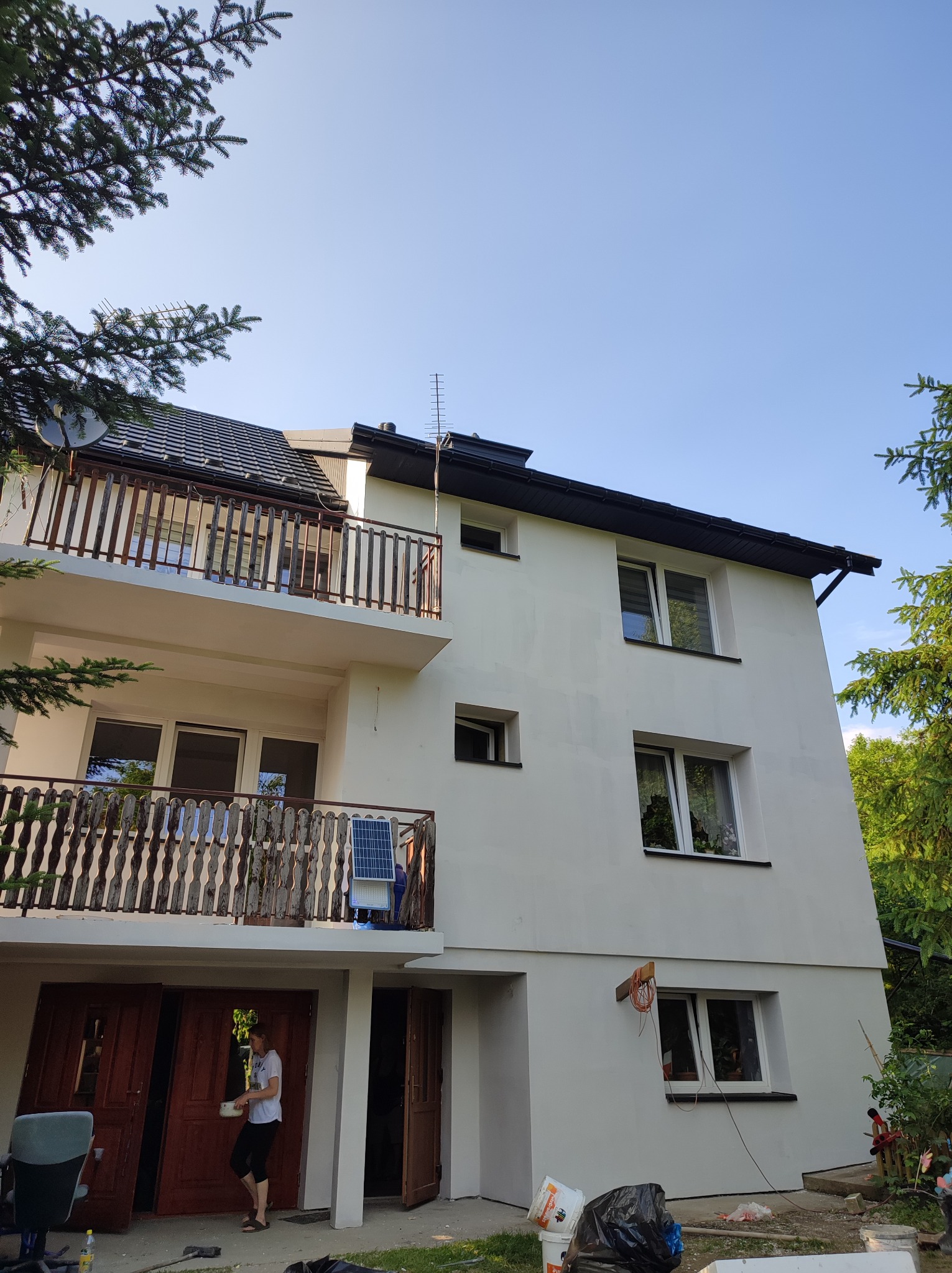 Biała elewacja dwupiętrowego domu z balkonami, widok z dołu. Osoba w pobliżu otwartych drzwi garażowych. Świeżo pomalowana fasada, widoczne ślady prac.