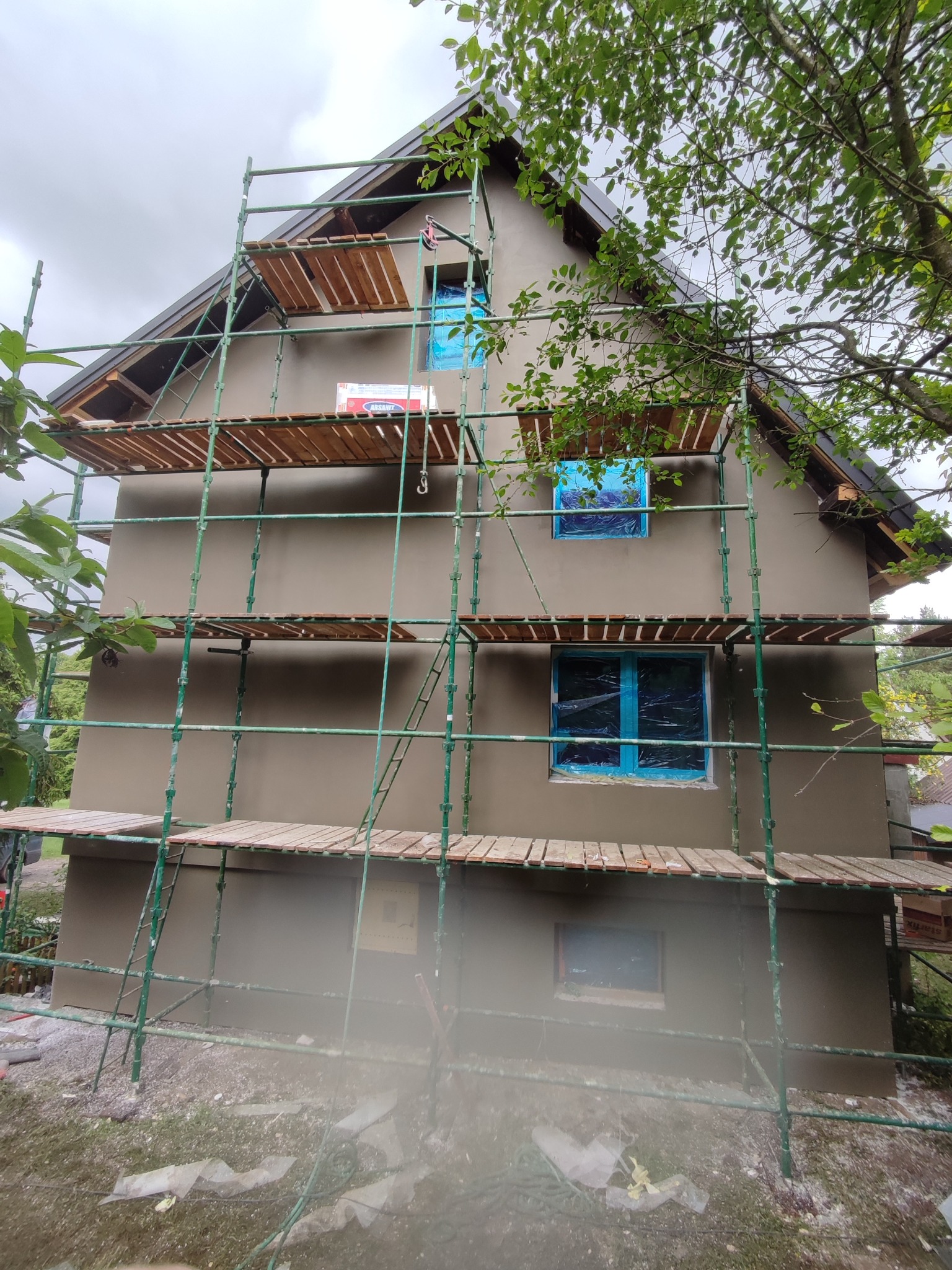 Dom jednorodzinny w trakcie prac elewacyjnych, z nałożonym tynkiem i rusztowaniem. Okna zabezpieczone folią. Widoczne detale wykończenia dachu i otoczenie budynku.