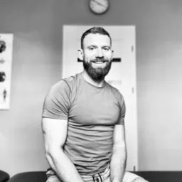 Uśmiechnięty mężczyzna z brodą siedzi na kozetce w gabinecie, w tle zegar i plakat anatomiczny. Zdjęcie czarno-białe.