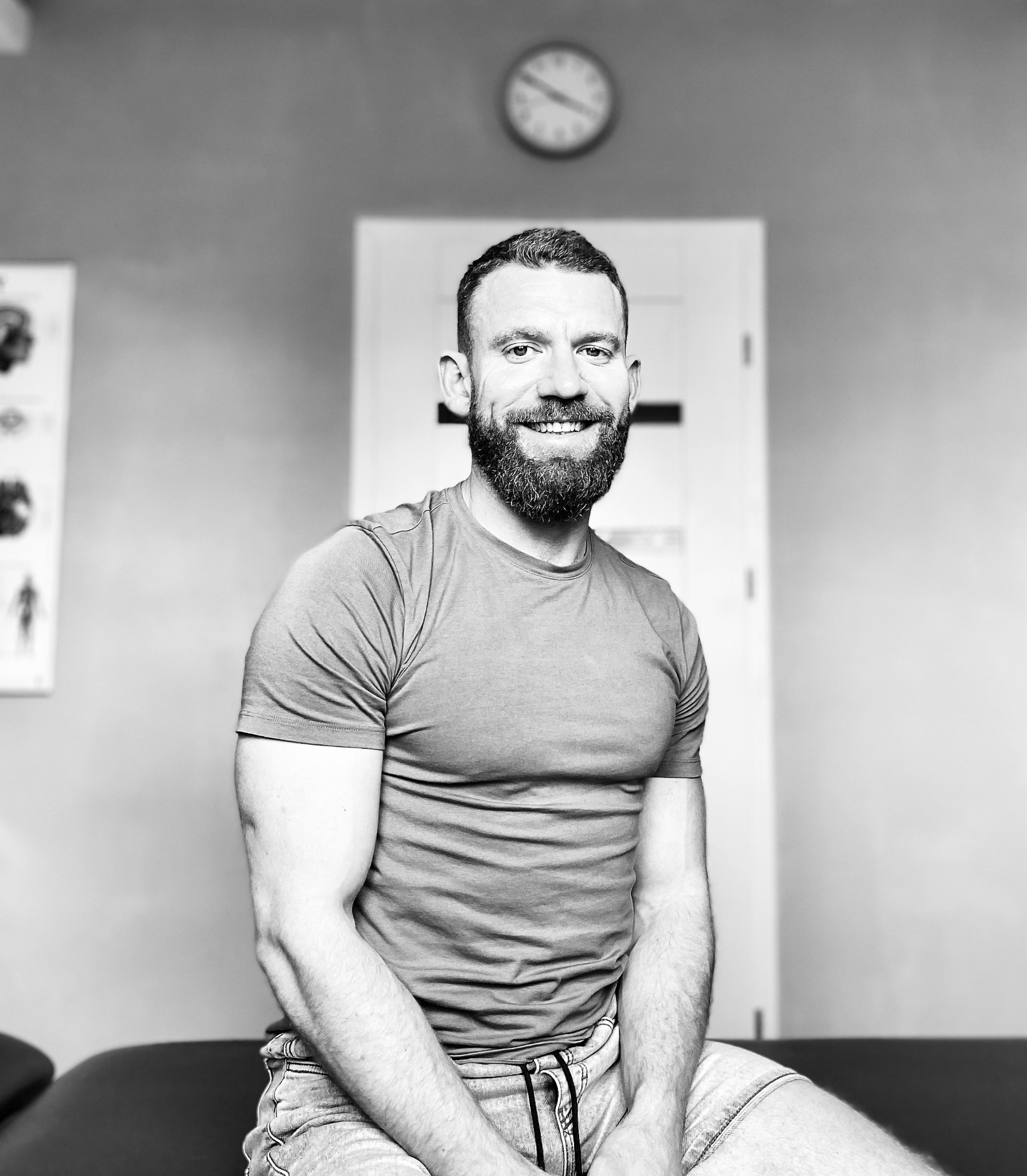 Uśmiechnięty mężczyzna z brodą siedzi na kozetce w gabinecie, w tle zegar i plakat anatomiczny. Zdjęcie czarno-białe.