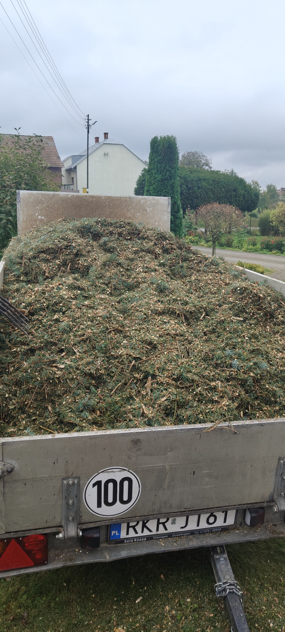 Przyczepa wypełniona po brzegi zrębkami drzewnymi, widoczna tablica rejestracyjna i znak ograniczenia prędkości do 100 km/h. W tle zabudowania i zieleń.