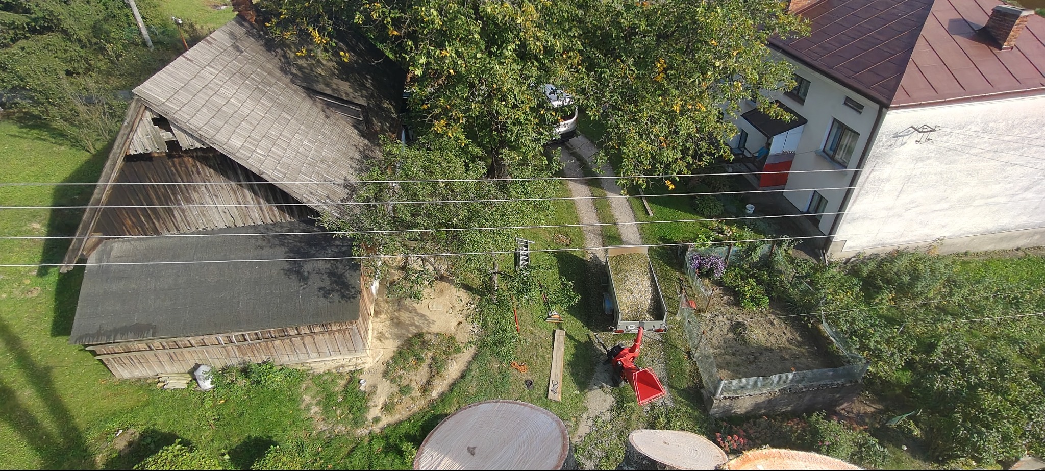 Widok z góry na posesję po wycince drzew: pnie, przyczepa z gałęziami, rozdrabniacz. Budynki w tle, zielony trawnik. Prace ogrodnicze w toku.