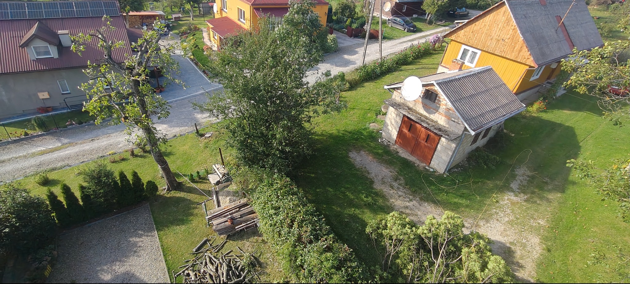 Widok z góry na posesję z drzewami, przyciętym żywopłotem i stertą gałęzi. W tle zabudowania, w tym garaż z anteną satelitarną.