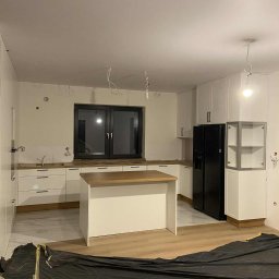 Meble na wymiar Wojciech Milewski - Wnętrze kuchni w trakcie montażu: białe szafki, drewniany blat, wyspa kuchenna. Na podłodze folia ochronna. Widoczne niedokończone instalacje elektryczne.