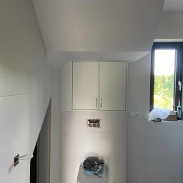 Meble na wymiar Wojciech Milewski - Biała łazienka w skosach z podwieszaną szafką nad toaletą. Widoczne płytki, okno i instalacja elektryczna w trakcie montażu. Podłoga imitująca marmur.