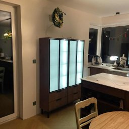 Meble na wymiar Wojciech Milewski - Nowoczesna szafa z podświetlanymi frontami w salonie, drewniane wykończenie, widoczny wieniec świąteczny, parkiet i fragment stołu z krzesłem.