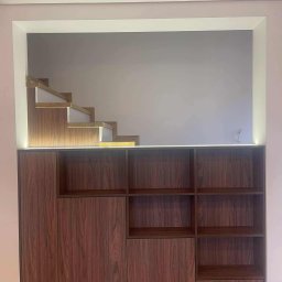 Meble na wymiar Wojciech Milewski - Nowoczesna szafa z drewna w kolorze orzecha, wbudowana we wnękę z podświetleniem LED, z widocznymi fragmentami schodów w górnej części. Minimalistyczny design.