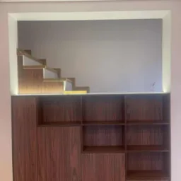 Nowoczesna szafa z drewna w kolorze orzecha, wbudowana we wnękę z podświetleniem LED, z widocznymi fragmentami schod&oacute;w w g&oacute;rnej części. Minimalistyczny design.